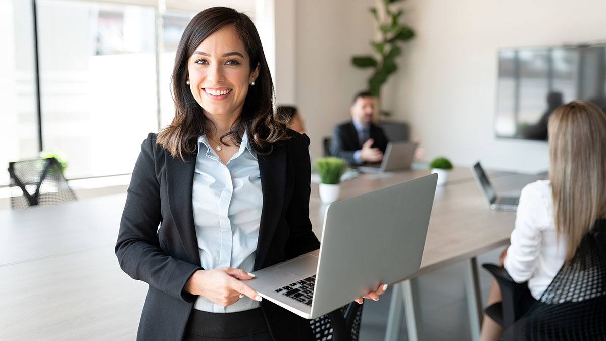 Smiling female holding a laptop in a conference room with a team in the background
