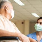 Masked female nurse kneeling down and talking to a masked, mature, male patient sitting in a wheelchair in a hospital
