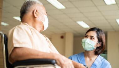 Masked female nurse kneeling down and talking to a masked, mature, male patient sitting in a wheelchair in a hospital