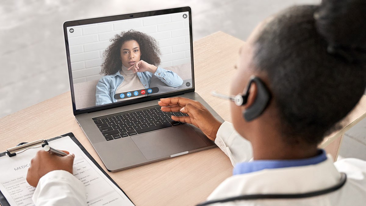 Clinician taking notes on clipboard while conducting a digital consultation with patient on computer