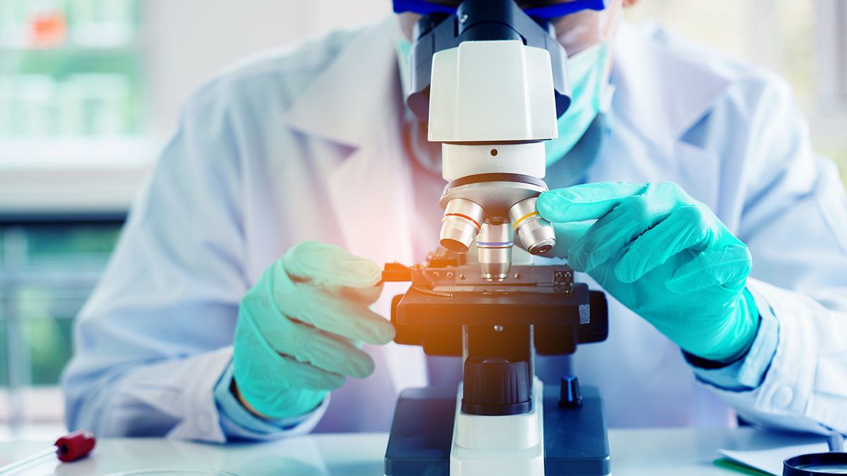 Scientist, wearing gloves and a mask, looks into a microscope