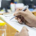 Hand holding silver pen and writing on a piece of paper attached to a clipboard
