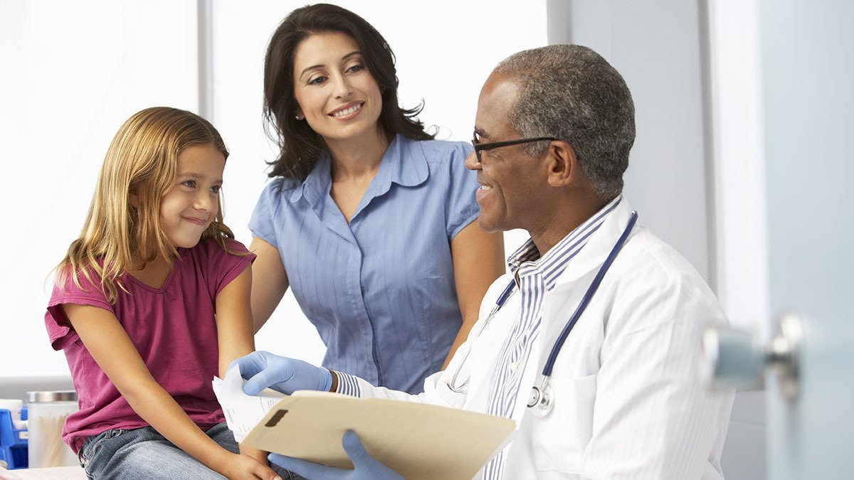 Older male doctor examining young girl patient, while her mom listens in, smiling